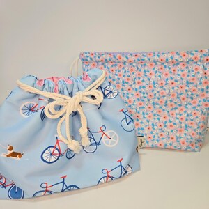 May include: Two drawstring bags. One is light blue with a bicycle and dog pattern, tied with a white rope. The other is a floral pattern with pink and orange flowers on a light blue background.