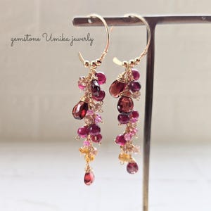 Peut inclure: Boucles d'oreilles créoles dorées ornées de grappes de pierres précieuses rouges et roses. Les boucles d'oreilles présentent une cascade de pierres de différentes tailles, dont des formes de goutte et rondes, suspendues aux créoles dorées. Le texte "gemstone Umika jewerly" est en haut à gauche.