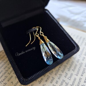 Puede incluir: Un par de pendientes colgantes con piedras facetadas de color azul claro en forma de lágrima, suspendidas de ganchos de alambre dorado. Las piedras están envueltas en alambre dorado en la parte superior. El texto "Umika accessory" está en blanco.