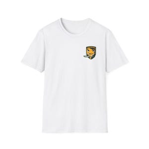 Può includere: T-shirt bianca con una piccola toppa colorata sulla parte superiore sinistra del petto. La toppa raffigura una testa di volpe con un coltello, su uno scudo verde e oro, con la scritta "FOX HOUND" sopra. La maglietta ha maniche corte e girocollo.