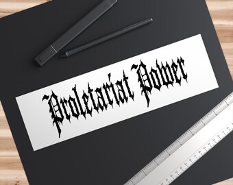 May include: A white sticker with the words "Proletariat Power" in a gothic font. The sticker is on a black surface, with a ruler and two black pens nearby. The sticker is rectangular.