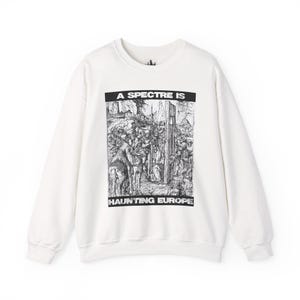 May include: A white sweatshirt with long sleeves featuring a black and white graphic print. The graphic depicts a historical scene with the text "A SPECTRE IS HAUNTING EUROPE" in a bold, black font. The sweatshirt has a crew neck.