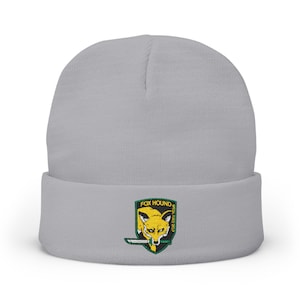 May include: A light gray beanie with a folded brim. The beanie features an embroidered patch with a yellow fox, a knife, and the text "FOX HOUND SPECIAL FORCE GROUP" in a green and black shield design.