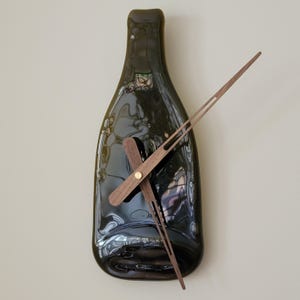 May include: A wall clock shaped like a dark green wine bottle. The clock hands are made of wood and are a light brown color. The clock is mounted on a light beige wall.