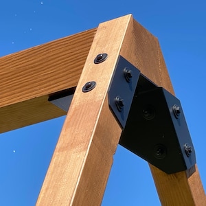 May include: A close-up of a wooden structure's corner, showing light brown wood and black metal brackets. The wood grain is visible, and the brackets are secured with black bolts. The background is a clear blue sky.