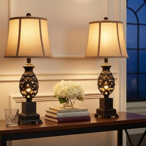 May include: Two matching table lamps with beige shades and ornate dark bases. The lamps sit on a dark wooden table, alongside a glass, books, and a vase of white flowers. The lamps are lit, casting a warm glow.
