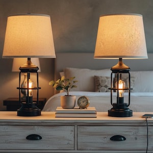 May include: Two matching black and gold table lamps with beige fabric shades. Each lamp features a caged design around a visible Edison-style bulb. The lamps sit on a white wooden nightstand, with a small plant, books, and a clock.