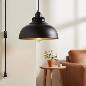 May include: Black pendant light with a gold interior, hanging from a black cord. The light is dome-shaped and has a classic, industrial design. A small plant in a terracotta pot sits on a wooden side table.
