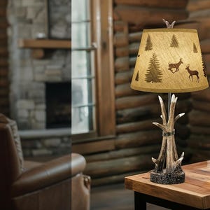 May include: A rustic table lamp with a deer antler base and a beige lampshade. The lampshade features a pattern of pine trees and deer silhouettes. The lamp is placed on a wooden table in a cabin setting.