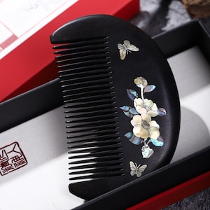 Puede incluir: Un peine de madera negra con mango curvo, adornado con flores y mariposas incrustadas de nácar. El peine se presenta en una caja negra con ribete rojo, lo que sugiere un juego de regalo. El diseño es elegante y decorativo.