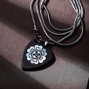 May include: A silver chain necklace with a black, triangular pendant. The pendant features a flower design with iridescent mother-of-pearl inlay. The necklace is displayed on a brown surface.