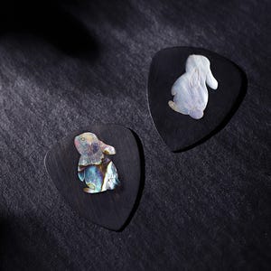 Puede incluir: Dos púas de guitarra negras con un diseño de conejo incrustado con nácar iridiscente. Las púas son triangulares y descansan sobre una superficie oscura y texturizada. Las figuras de conejos tienen detalles.