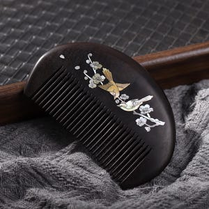 Puede incluir: Un peine de madera marrón oscuro con dientes finos. El peine está decorado con un diseño incrustado de dos pájaros y ramas florecientes en tonos dorados, blancos y plateados. El peine está sobre una tela gris.