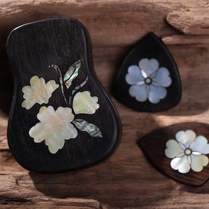 May include: A black wooden guitar pick holder and three guitar picks. The holder and picks are decorated with mother-of-pearl flower designs. The background is a textured, wooden surface.