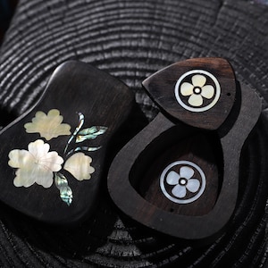 May include: Two dark wooden boxes with floral designs. One box is open, revealing a flower-shaped inlay. The other box features a mother-of-pearl floral design. The boxes are set against a dark, textured background.