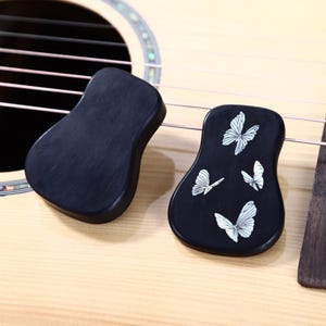 May include: Two black guitar-shaped accessories, one plain and one decorated with white butterfly designs, rest on a light-colored acoustic guitar. The guitar has strings and a dark wooden bridge.
