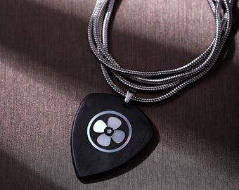 May include: A black pendant necklace with a silver chain. The pendant is a dark, guitar pick shape with a white flower design in the center. The necklace is displayed on a brown surface.