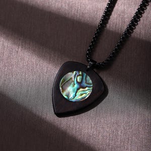 Puede incluir: Un collar con colgante de madera oscura con forma triangular y una incrustación circular de concha de abulón. El collar tiene una cadena negra. La concha de abulón tiene colores iridiscentes azules, verdes y dorados.