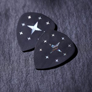 May include: Two black guitar picks with a star and small star accents. The large star is white and the small stars are white. The picks are on a dark gray surface.