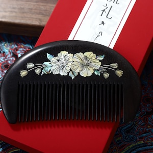 Puede incluir: Un peine de madera negra con mango curvo, adornado con un diseño floral incrustado con nácar. El peine descansa sobre una caja roja, lo que sugiere un artículo de regalo. El diseño floral presenta acentos blancos y verde claro.