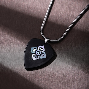 May include: A black pendant necklace with a silver chain. The pendant is a dark, triangular shape with a floral design inlaid with iridescent mother-of-pearl. The necklace is displayed on a textured, brown surface.