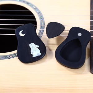 May include: Acoustic guitar with three black guitar picks. Two picks are black with a curved shape, one featuring a white moon and a bunny design. The third pick is a standard triangle shape.