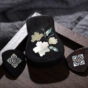 May include: A dark wooden guitar pick holder and two guitar picks with inlaid mother-of-pearl floral designs. The holder features a larger floral design. The items are displayed on a wooden tray.