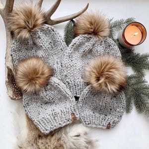 May include: Several grey knitted hats, each with a large, fluffy brown pom-pom. The hats are displayed on a white surface with a lit candle, deer antlers, and fir branches. The hats appear to be hand-knitted.