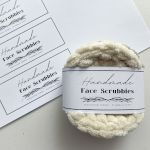 May include: A stack of handmade face scrubbies with a white label wrapped around them. The label reads "Handmade Face Scrubbies" with a leaf design. The scrubbies are a light beige color and are next to a stack of labels.