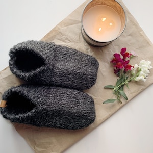 May include: A pair of dark gray knitted slippers with a small leather tag, resting on brown paper. A lit candle in a metal container and a small bouquet of flowers are also present, creating a cozy, homey scene.
