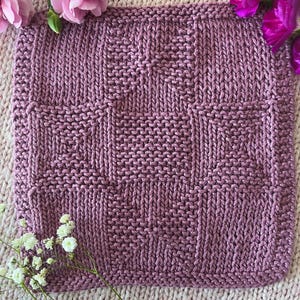 May include: A square, hand-knitted item in a mauve color, featuring a textured pattern of vertical and horizontal stitches. The item is surrounded by pink flowers and white baby's breath, creating a soft, feminine aesthetic. The knitted piece is likely a washcloth or small blanket.