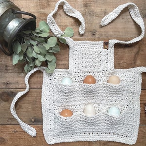 May include: A cream-colored, crocheted egg-gathering apron with six pockets, holding various colored eggs. The apron has a neck strap and waist ties. A metal container with eucalyptus sprigs is in the background, set on a wooden surface.