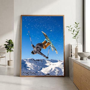 May include: A framed wall art print featuring a skier performing an aerial jump against a clear blue sky. The skier wears a dark patterned jacket and orange pants, holding ski poles while suspended above snow-covered mountain peaks.