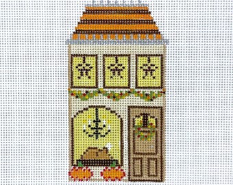 May include: A cross-stitch pattern of a house with a brown roof and tan walls. The house has three windows with yellow interiors and a door with a wreath. Pumpkins are at the base of the house.