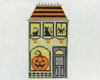 May include: A needlepoint Halloween house with a black and orange striped roof, three windows with black cat and spider designs, and a jack-o'-lantern. The house has a green facade, a gray door, and a small fence.