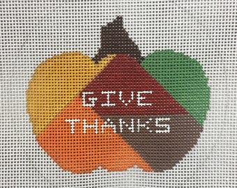May include: A needlepoint canvas with a pumpkin design. The pumpkin is made up of different colored sections, including green, brown, red, orange, and yellow. The words "GIVE THANKS" are stitched in white on the pumpkin.
