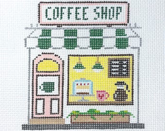 May include: A cross-stitch pattern on white fabric depicting a coffee shop storefront. The design features a pink door, a yellow window displaying a cake and coffee pot, a green striped awning, and the text COFFEE SHOP in dark green.