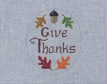 May include: A white canvas with a brown inscription that reads "Give Thanks". The inscription is surrounded by a circle of colorful leaves and an acorn.