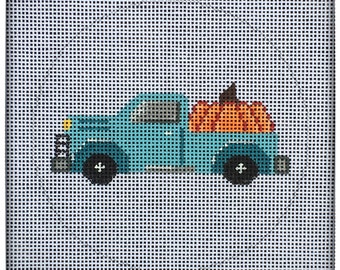 May include: A needlepoint canvas featuring a light blue pickup truck carrying a large orange pumpkin in its bed. The truck has black wheels, a yellow headlight, and a brown stem on the pumpkin, all set against a white grid background.