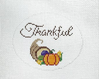 May include: A white canvas with the word "Thanksgiving" stitched in brown thread. A cornucopia with a brown, orange, and purple design is stitched below the word. The cornucopia is filled with a variety of fruits and vegetables, including a pumpkin, grapes, and leaves.