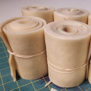 May include: Six rolls of beige material, each secured with a rubber band. The rolls are tightly wound, displaying a spiral pattern on their tops. A piece of paper with the numbers "48-52" is visible.