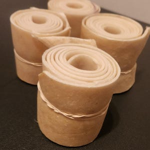 May include: Four rolls of light brown material, possibly leather or paper, are arranged on a dark surface. Each roll is tightly wound, revealing concentric circles. A thin band secures each roll.
