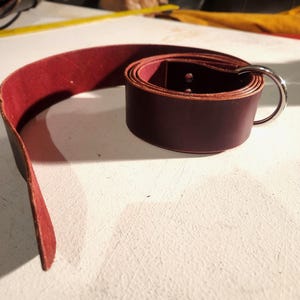 May include: A rolled burgundy leather belt with a silver metal ring buckle. The belt is unrolled on a white surface, showing its full length. The leather appears to be thick and durable, with a rich, deep color.