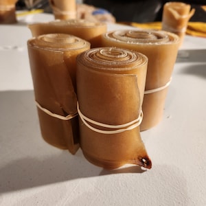 May include: Several rolls of brown, rolled-up material, possibly food items, are bundled together with white rubber bands. The rolls are tightly wound, displaying a spiral pattern on their tops. The items are on a white surface.