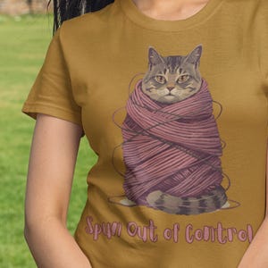 May include: Mustard-coloured t-shirt with a cat illustration wrapped in pink yarn. The cat has grey and brown fur and is looking at the viewer. The text "Spun Out of Control" is printed in a pink script font below the cat.