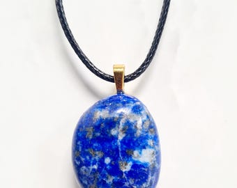 以下が含まれることがあります： 青いラピスラズリのペンダントネックレス。 楕円形の石は、金と白の斑点のある深い青色です。 金色の金属製のベイルが付いた黒いコードネックレスに取り付けられています。