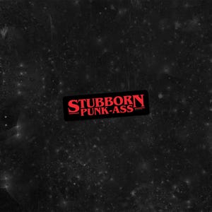 May include: A black rectangular sticker with the words "STUBBORN PUNK-ASS" in red, stylized lettering. The sticker has a black border and is set against a dark, speckled background.