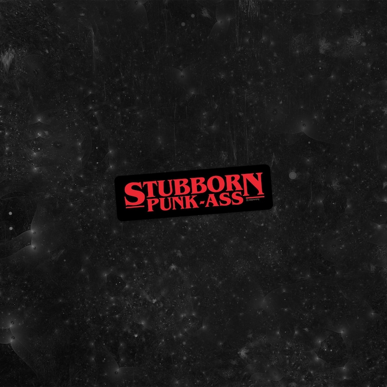 May include: A black rectangular sticker with the words "STUBBORN PUNK-ASS" in red, stylized lettering. The sticker has a black border and is set against a dark, speckled background.