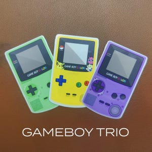 May include: Three Game Boy Color handheld game consoles in green, yellow, and purple. The yellow console features Pikachu and other characters. Each console has a screen, buttons, and the text "GAME BOY COLOR". The image also includes the text "GAMEBOY TRIO".