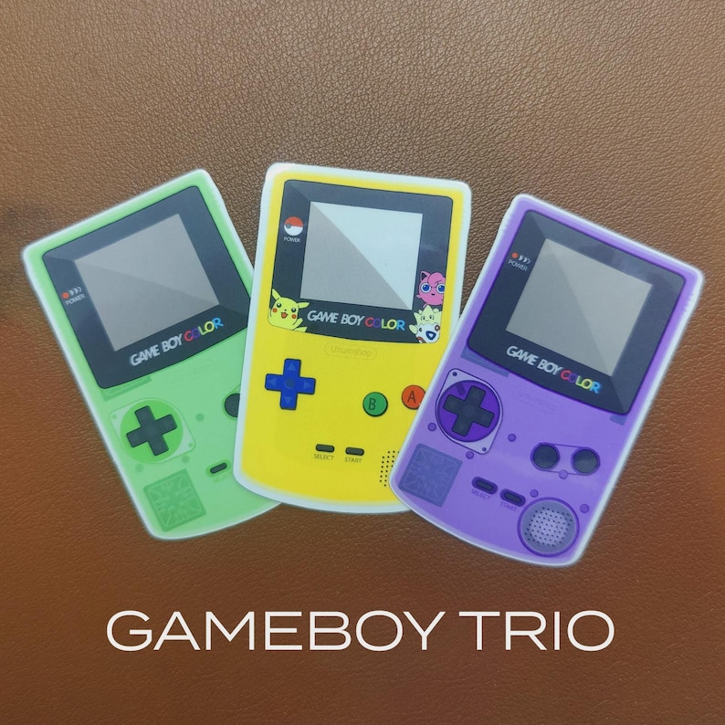 May include: Three Game Boy Color handheld game consoles in green, yellow, and purple. The yellow console features Pikachu and other characters. Each console has a screen, buttons, and the text "GAME BOY COLOR". The image also includes the text "GAMEBOY TRIO".